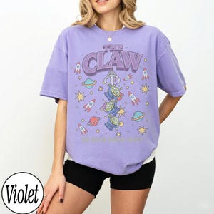 Puede incluir: Una camiseta violeta de manga corta con un estampado gr&aacute;fico de tres alien&iacute;genas verdes colgados del gancho de una m&aacute;quina de garra. El dise&ntilde;o incluye el texto THE CLAW, rodeado de cohetes, planetas y estrellas coloridas en el frente.