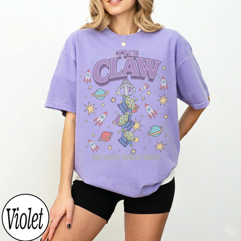 Puede incluir: Una camiseta violeta de manga corta con un estampado gr&aacute;fico de tres alien&iacute;genas verdes colgados del gancho de una m&aacute;quina de garra. El dise&ntilde;o incluye el texto THE CLAW, rodeado de cohetes, planetas y estrellas coloridas en el frente.