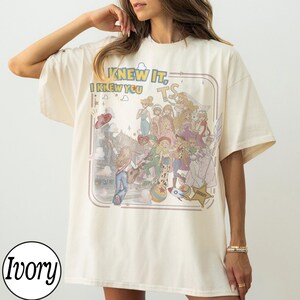 May include: An ivory short-sleeved t-shirt featuring a graphic print of various animated characters and the text "KNEW IT, I KNEW YOU" and "TS". The design includes a star-shaped sheriff badge, a rocket, and a ball, all set within a rounded frame.