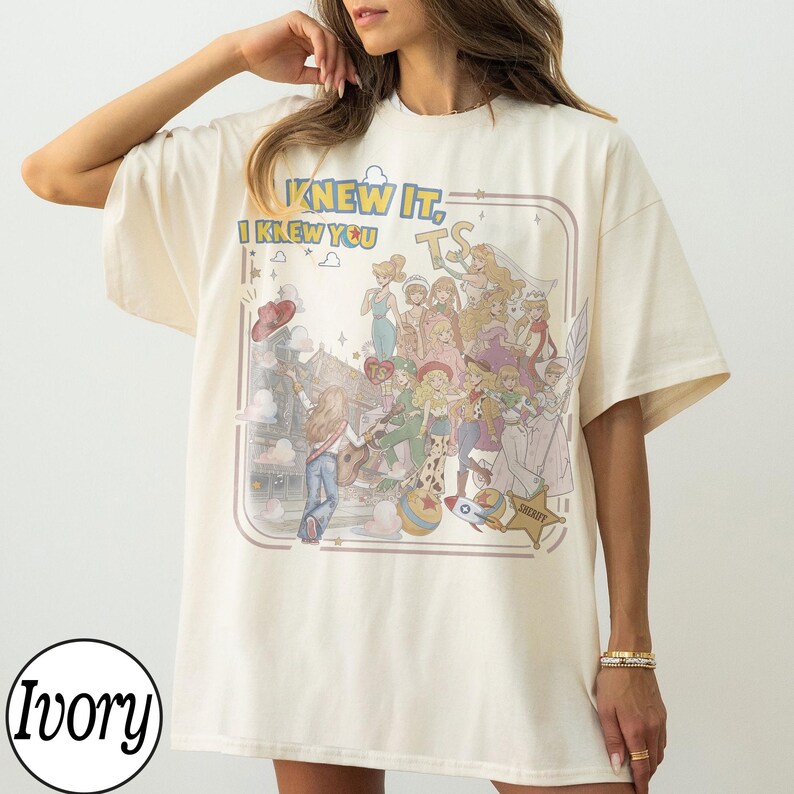 May include: An ivory short-sleeved t-shirt featuring a graphic print of various animated characters and the text "KNEW IT, I KNEW YOU" and "TS". The design includes a star-shaped sheriff badge, a rocket, and a ball, all set within a rounded frame.