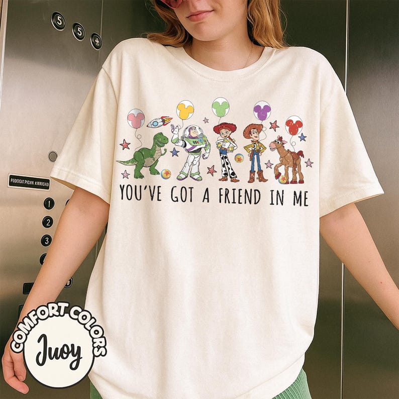 May include: Cream-colored t-shirt featuring a colorful graphic of cartoon characters and balloons. The text "YOU'VE GOT A FRIEND IN ME" is printed below the characters. The shirt has short sleeves and a crew neck.