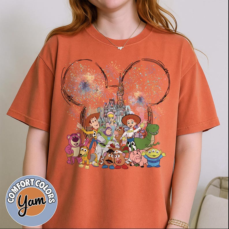 以下が含まれることがあります： オレンジ色のコンフォートカラーズTシャツ。ディズニーをテーマにしたデザインで、お城、花火、トイ・ストーリーのキャラクターが描かれています。丸首で半袖。ブランドロゴが左下に表示されています。