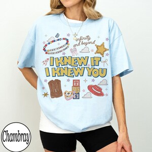 May include: A light blue t-shirt featuring a graphic print with the text "I KNEW IT I KNEW YOU" and "infinity and beyond". The design includes illustrations of a beaded bracelet, a gold star, cowboy boots, alphabet blocks, and a red hat.