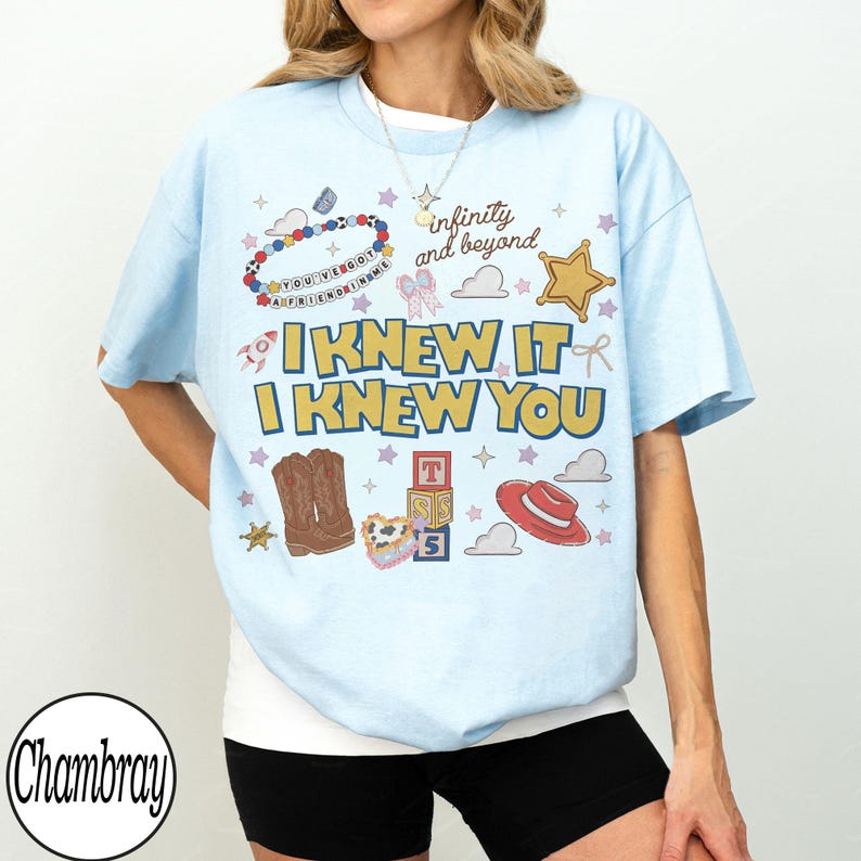 May include: A light blue t-shirt featuring a graphic print with the text "I KNEW IT I KNEW YOU" and "infinity and beyond". The design includes illustrations of a beaded bracelet, a gold star, cowboy boots, alphabet blocks, and a red hat.