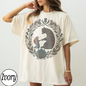 以下が含まれることがあります： アイボリー色のTシャツ。円形のデザインで、クマと女の子が手をつないでおり、花のモチーフと動物のモチーフで囲まれています。クマと女の子は青、茶色、グレーの色合いです。Tシャツは半袖で、ゆったりとしたフィット感です。