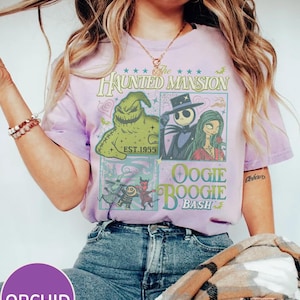 May include: Orchid purple t-shirt featuring a graphic design with characters from The Haunted Mansion and the text "Oogie Boogie Bash". The design includes images of Jack Skellington, Sally, and Oogie Boogie. The shirt is worn with blue jeans.