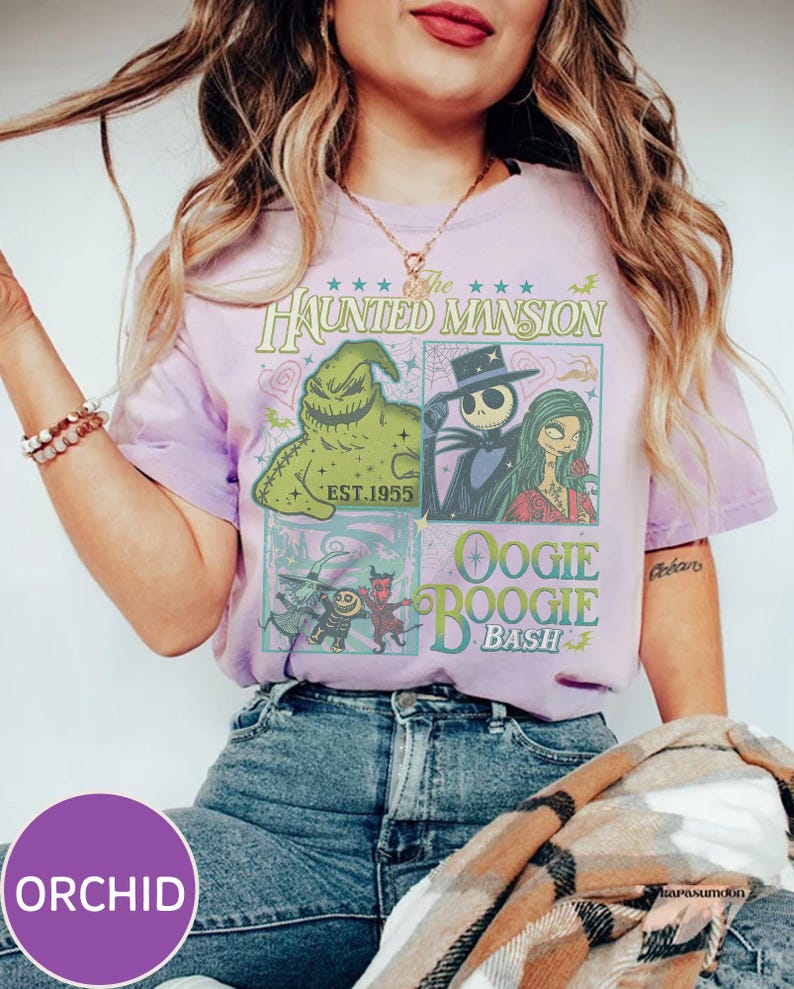 May include: Orchid purple t-shirt featuring a graphic design with characters from The Haunted Mansion and the text "Oogie Boogie Bash". The design includes images of Jack Skellington, Sally, and Oogie Boogie. The shirt is worn with blue jeans.