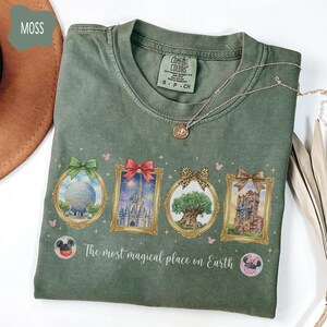 K&ouml;nnte beinhalten: Moosgr&uuml;nes T-Shirt mit vier gerahmten Bildern mit Schleifen. Die Bilder zeigen eine Kugel, ein Schloss mit Feuerwerk, einen Baum und einen Turm. Der Text "The most magical place on Earth" ist darunter gedruckt.