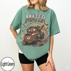 May include: Light green t-shirt featuring a graphic of Mater from the movie Cars. The word "MATER" is printed across the top. The cartoon tow truck is brown with a smiling face and is surrounded by tools and lightning bolts.