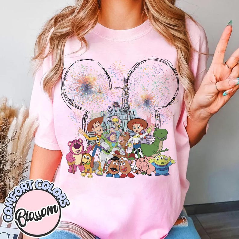 以下が含まれることがあります： ピンク色のTシャツ。ディズニーのトイ・ストーリーのキャラクター、花火、お城のグラフィックが描かれ、ミッキーマウスの耳で縁取られています。「COMFORT COLORS Blossom」の文字が円形デザインで含まれています。
