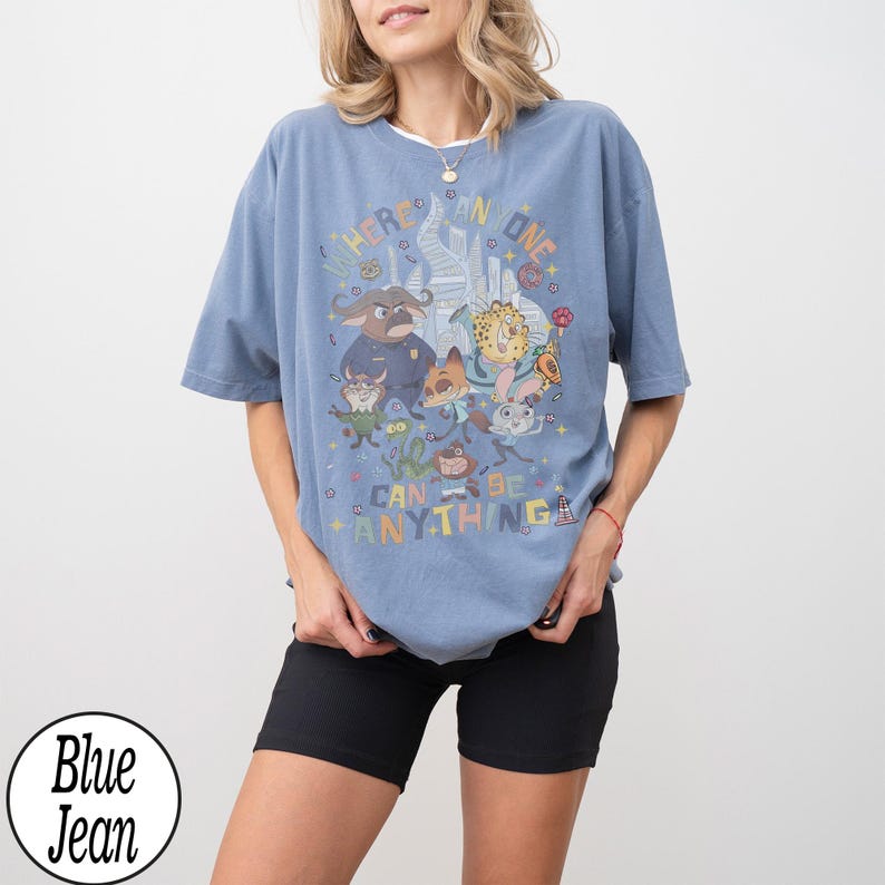 May include: Light blue t-shirt with a cartoon graphic and the words "Where Anyone Can Be Anything." The design features various cartoon characters. The shirt has a relaxed fit.
