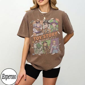 May include: A brown short-sleeve t-shirt featuring a graphic print of Toy Story characters in Halloween costumes. The design includes Buzz Lightyear, Woody, Jessie, Rex, Slinky Dog, and Bo Peep with the text Toy Story Est. 1995 and spiderwebs.