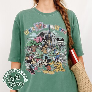May include: Light green Comfort Colors t-shirt featuring a colorful graphic of Disney characters and the text "Walt Disney World". The design includes Mickey Mouse, Minnie Mouse, Donald Duck, and other characters in front of a castle.