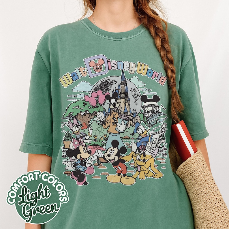 May include: Light green Comfort Colors t-shirt featuring a colorful graphic of Disney characters and the text "Walt Disney World". The design includes Mickey Mouse, Minnie Mouse, Donald Duck, and other characters in front of a castle.