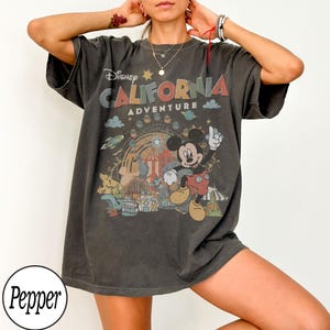 May include: Dark gray t-shirt featuring a colorful graphic of Mickey Mouse in front of a Ferris wheel and the words "Disney California Adventure". The shirt has short sleeves and a relaxed fit, perfect for casual wear.
