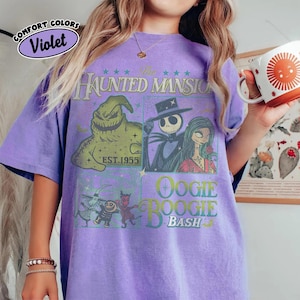 May include: Violet Comfort Colors t-shirt featuring a graphic design from The Haunted Mansion, with characters Jack Skellington, Sally, and Oogie Boogie. The shirt has a vintage look with the text "EST. 1955" and "Oogie Boogie Bash".
