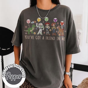 May include: Dark gray t-shirt featuring characters from the movie Toy Story, including Rex, Buzz Lightyear, Woody, and Jessie. The shirt has the phrase "You've Got a Friend in Me" printed across the front. The shirt also has the Comfort Colors Pepper logo.