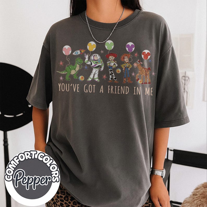 May include: Dark gray t-shirt featuring characters from the movie Toy Story, including Rex, Buzz Lightyear, Woody, and Jessie. The shirt has the phrase "You've Got a Friend in Me" printed across the front. The shirt also has the Comfort Colors Pepper logo.