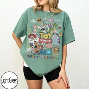 May include: A light green short-sleeve t-shirt featuring a colorful graphic print of Toy Story characters including Buzz Lightyear, Woody, and Jessie. The text reads "TOY STORY" and "YOU'VE GOT A FRIEND IN ME" in bold, multi-colored lettering.