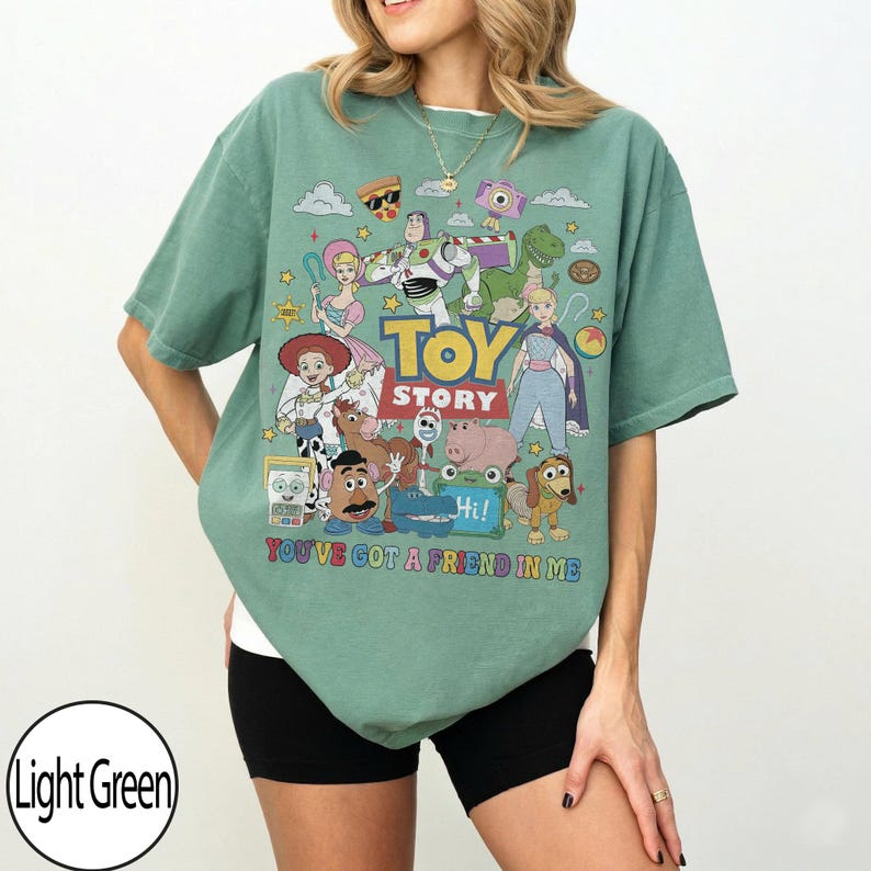May include: A light green short-sleeve t-shirt featuring a colorful graphic print of Toy Story characters including Buzz Lightyear, Woody, and Jessie. The text reads "TOY STORY" and "YOU'VE GOT A FRIEND IN ME" in bold, multi-colored lettering.