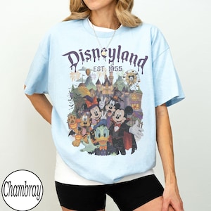 Puede incluir: Una camiseta azul claro con un estampado gr&aacute;fico de personajes de Disney con disfraces de Halloween, incluidos Mickey Mouse, Minnie Mouse, Goofy, el Pato Donald y Pluto. El texto dice Disneyland EST. 1955 sobre un castillo y una noria.
