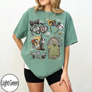 May include: Light green t-shirt featuring a colorful graphic of characters from a popular animated movie. The design includes Jack Skellington, Sally, and Oogie Boogie. The shirt is a relaxed fit and has short sleeves.