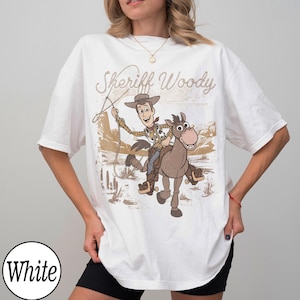 May include: White t-shirt featuring a graphic of Sheriff Woody riding a horse with the text "Sheriff Woody" in a vintage style. The design is in shades of brown, beige, and tan.