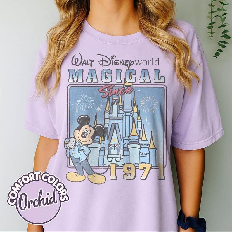 May include: A light purple t-shirt with a graphic featuring Mickey Mouse in front of Cinderella's castle. The text reads "Walt Disney World MAGICAL Since 1971". The shirt has a Comfort Colors Orchid label.