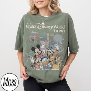 May include: A moss green short-sleeve t-shirt featuring a graphic print of Walt Disney World Est. 1971. The design includes illustrations of Mickey Mouse, Minnie Mouse, Donald Duck, Daisy Duck, Goofy, Pluto, Chip, and Dale in front of park landmarks.