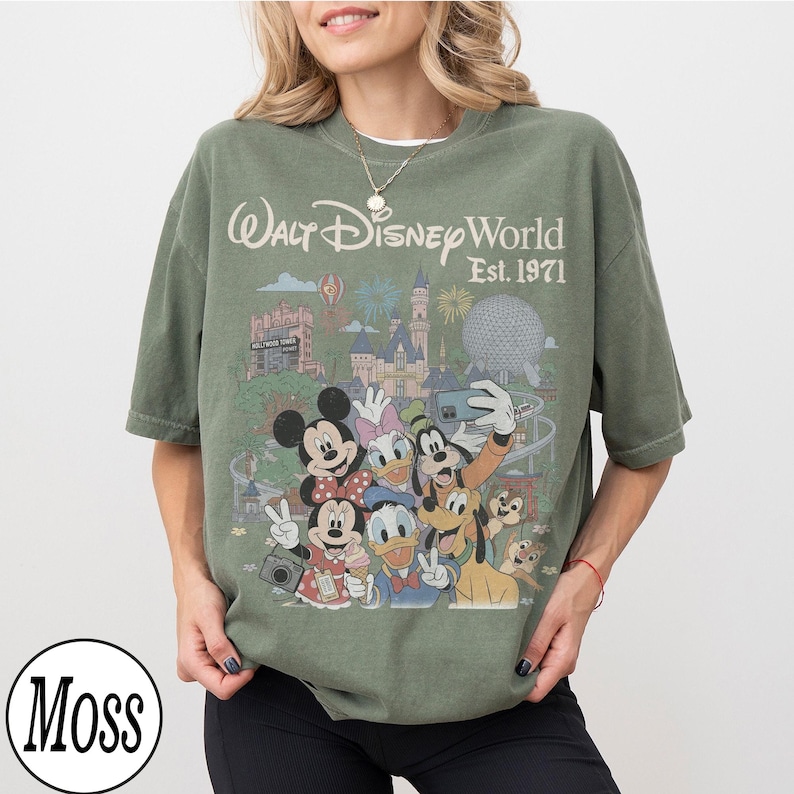 May include: A moss green short-sleeve t-shirt featuring a graphic print of Walt Disney World Est. 1971. The design includes illustrations of Mickey Mouse, Minnie Mouse, Donald Duck, Daisy Duck, Goofy, Pluto, Chip, and Dale in front of park landmarks.