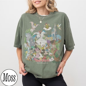 May include: Olive green t-shirt featuring a colorful graphic of Disney characters, including Mickey Mouse, Minnie Mouse, and Dumbo. The text "Magic Kingdom, Our Disney World" is printed below the characters. The shirt has short sleeves.