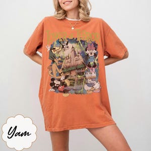 K&ouml;nnte beinhalten: Orangefarbenes T-Shirt mit einer Grafik von Disney-Figuren, darunter Micky Maus, Minnie Maus und Goofy, mit dem Text "Tower of Terror". Das Design umfasst ein Schloss und Blitze.