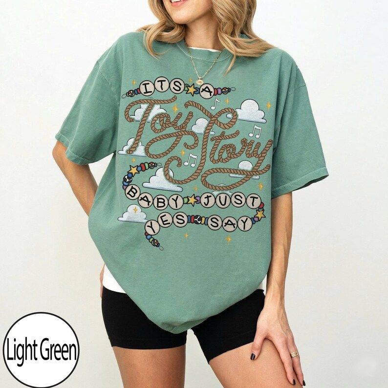 May include: A light green short-sleeve t-shirt featuring a graphic design with rope-style lettering that reads "ITS A Toy Story BABY JUST YES SAY". The print includes white clouds, small yellow stars, musical notes, and colorful beads.