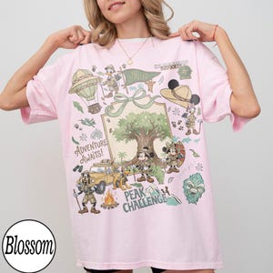 May include: Pale pink t-shirt with a vibrant graphic of cartoon characters, a hot air balloon, and the words "Adventure Awaits!". The design includes a tree, a jeep, and the words "Peak Challenge".