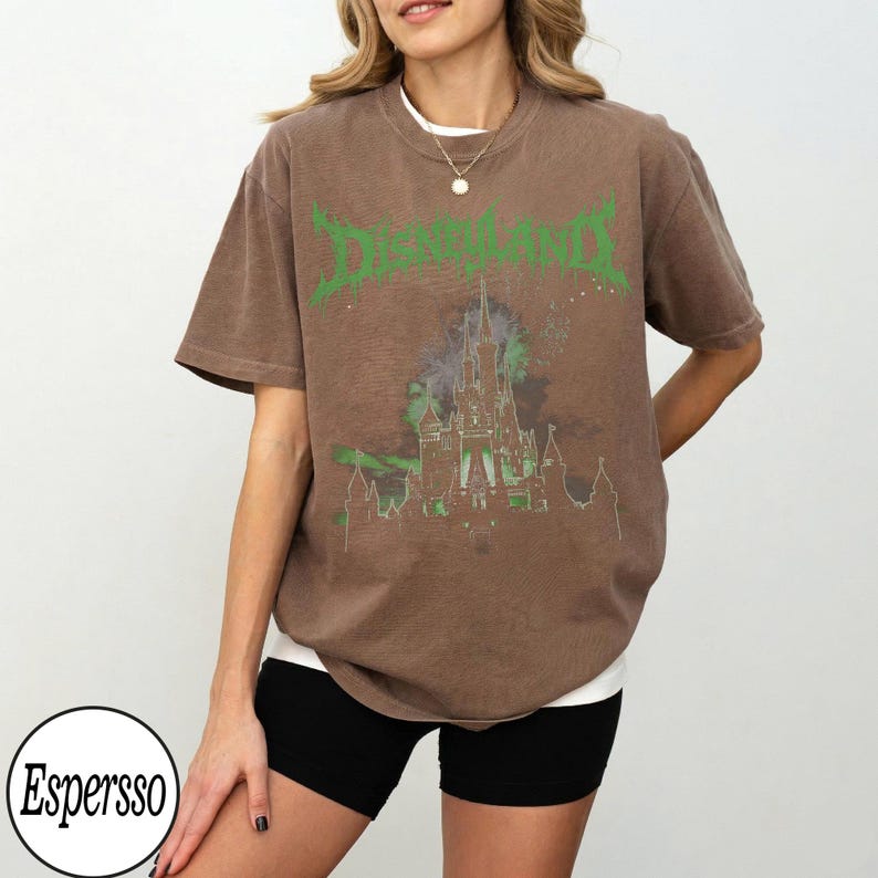 May include: Brown t-shirt with a green "Disneyland" graphic and a castle design. The shirt has a vintage, distressed look and a relaxed fit. The shirt is worn with black shorts and a white undershirt.
