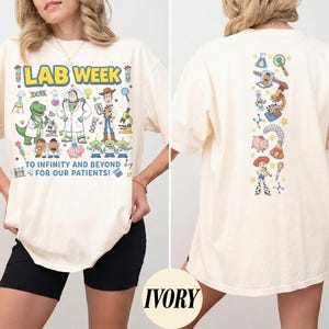 Op de afbeelding: Ivoor t-shirt met een kleurrijke afbeelding van Toy Story-personages. Op de voorkant staat "LAB WEEK" en "TO INFINITY AND BEYOND FOR OUR PATIENTS!". De achterkant heeft een verticaal ontwerp met meer Toy Story-personages.