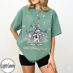 May include: Light green t-shirt with a graphic of Goofy in front of Cinderella's castle at Walt Disney World. The design includes fireworks and Mickey Mouse silhouettes. The text "Walt Disney World" is printed below the graphic.