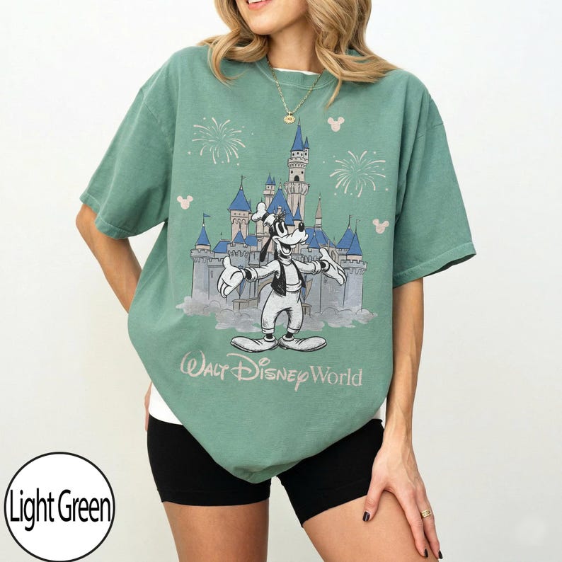 May include: Light green t-shirt with a graphic of Goofy in front of Cinderella's castle at Walt Disney World. The design includes fireworks and Mickey Mouse silhouettes. The text "Walt Disney World" is printed below the graphic.