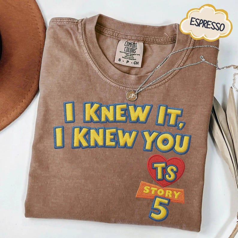 Puede incluir: Una camiseta marr&oacute;n de manga corta con el texto bordado I KNEW IT, I KNEW YOU en amarillo con contornos azules. Debajo del texto hay un coraz&oacute;n rojo con las letras TS, la palabra STORY y el n&uacute;mero 5 en amarillo con contornos azules.