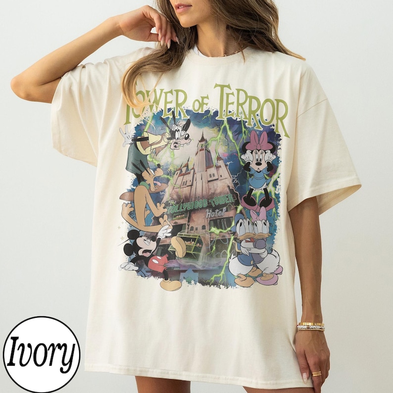 K&ouml;nnte beinhalten: Elfenbeinfarbenes T-Shirt mit einem Aufdruck von Mickey Mouse, Goofy, Minnie Mouse und Donald Duck vor dem Hollywood Tower Hotel. Der Text "Tower of Terror" ist in Gr&uuml;n &uuml;ber dem Aufdruck gedruckt. Das Shirt hat kurze &Auml;rmel.
