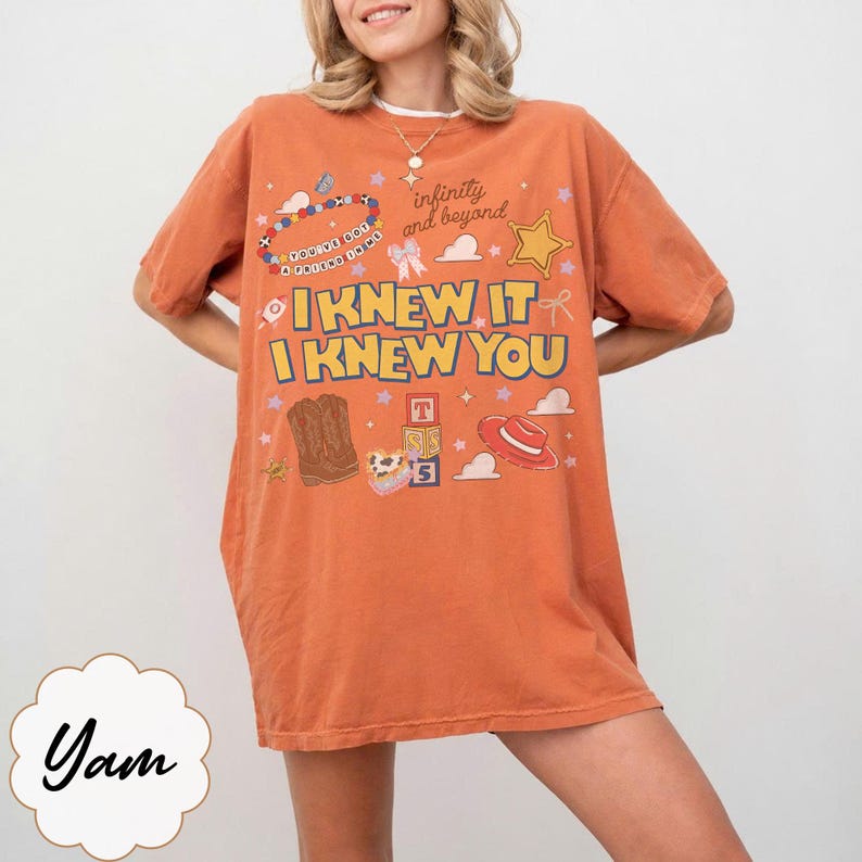 May include: An orange short-sleeve t-shirt featuring a graphic print with the text "I KNEW IT I KNEW YOU" and "infinity and beyond". The design includes illustrations of a beaded necklace, a star, cowboy boots, alphabet blocks, and a red hat.