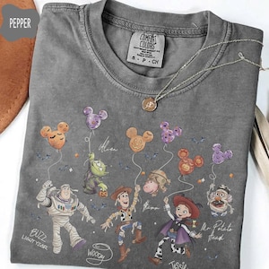 Pode incluir: Uma camiseta cinza Comfort Colors com os personagens da Disney Buzz Lightyear, Woody, Jessie e Sr. Batata. Os personagens est&atilde;o segurando bal&otilde;es em forma de Mickey Mouse. A camisa tamb&eacute;m tem o texto "BUZZ LIGHT YEAR", "WOODY", "Jessie" e "Mr Potato Head". Um colar de prata est&aacute; sobre a camisa.