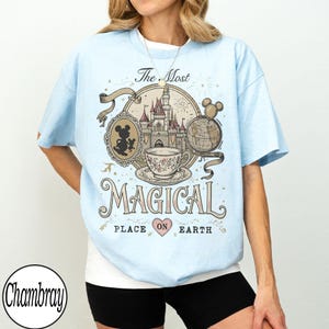 May include: Light blue t-shirt featuring a whimsical design with a castle, teacup, and globe. The text reads "The Most Magical Place on Earth." A silhouette of Mickey Mouse is also included in the design.