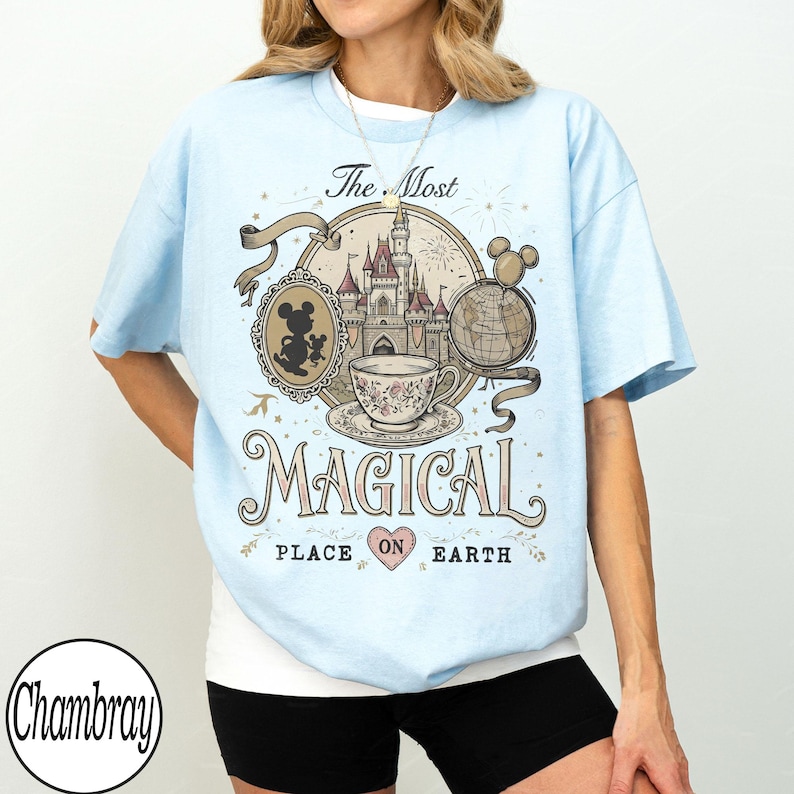 May include: Light blue t-shirt featuring a whimsical design with a castle, teacup, and globe. The text reads "The Most Magical Place on Earth." A silhouette of Mickey Mouse is also included in the design.