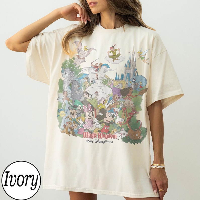 May include: Ivory-colored t-shirt featuring a colorful graphic of various Disney characters, including Mickey Mouse, Minnie Mouse, and Cinderella's castle. The text "Magic Kingdom Walt Disney World" is printed below the graphic.