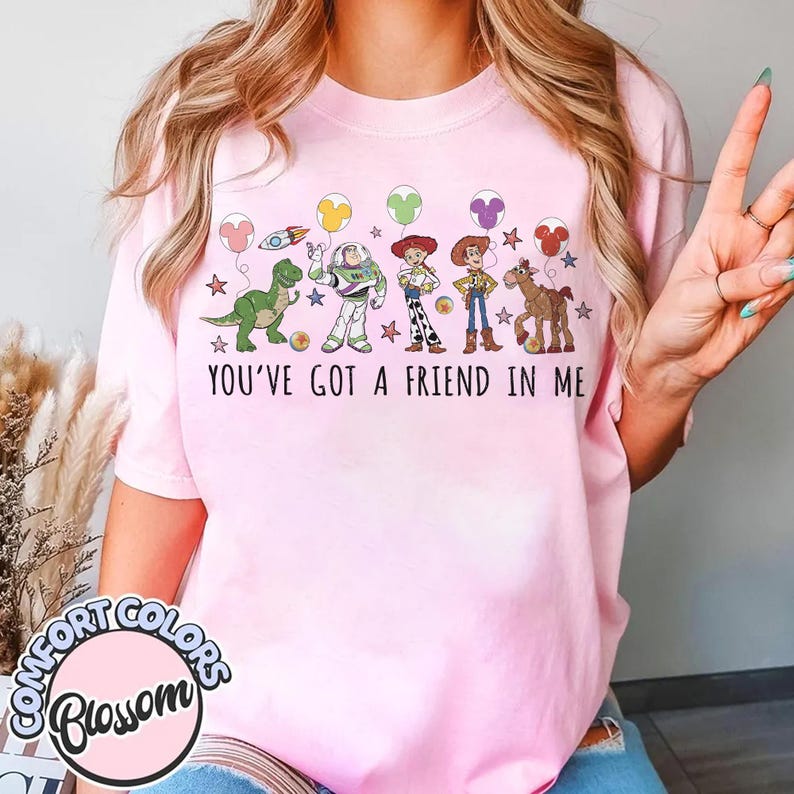 May include: Pink t-shirt featuring a graphic of Toy Story characters, including Rex, Buzz Lightyear, Woody, and Bullseye, with the text "YOU'VE GOT A FRIEND IN ME." The design includes balloons and stars. The shirt has a Comfort Colors Blossom logo.