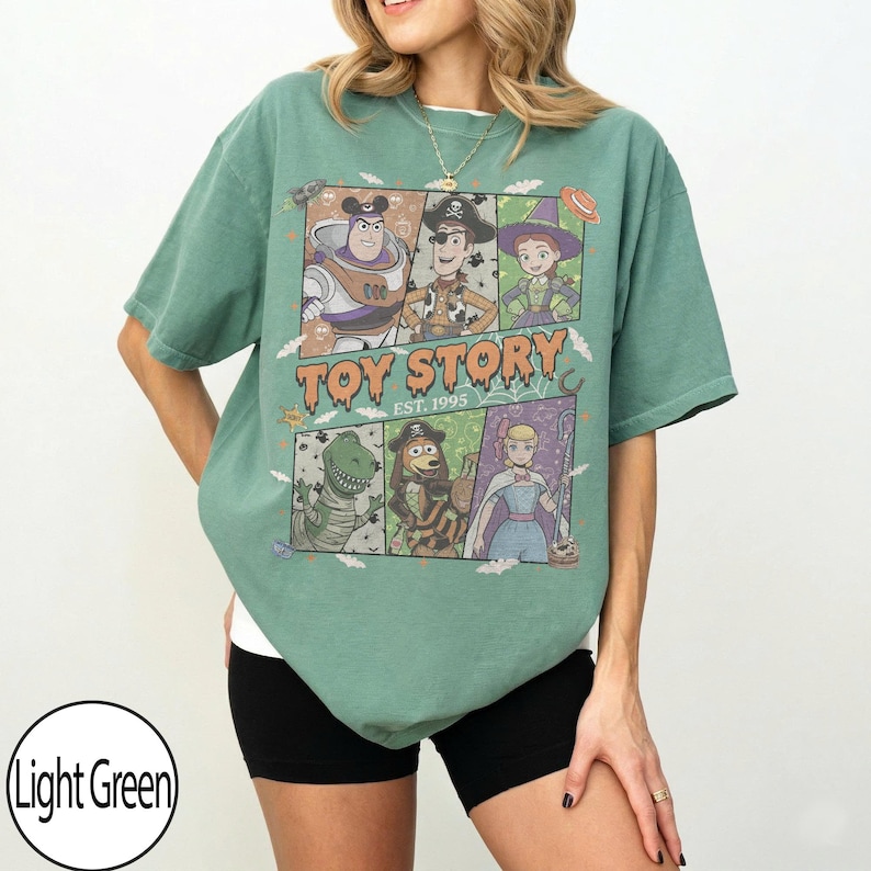 May include: A light green short-sleeve t-shirt featuring a graphic print of Toy Story characters in Halloween costumes. The text reads Toy Story Est. 1995. The design includes Buzz Lightyear, Woody, Jessie, Rex, Slinky Dog, and Bo Peep in panels.