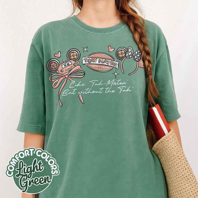 Puede incluir: Camiseta verde claro Comfort Colors con un gr&aacute;fico de Tow Mater de Cars. El dise&ntilde;o incluye un lazo rosa, ruedas de coche y el texto "Tow Mater Towing & Salvage" y "Like Tuh-Mater But without the Tuh."