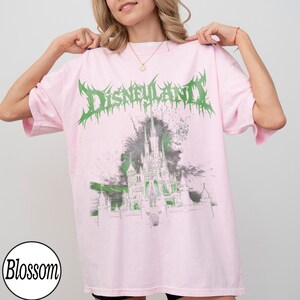 May include: Pink t-shirt with a green and white graphic of a castle and the word "Disneyland" in a spiky font. The shirt is a light pink color and has a round neckline.