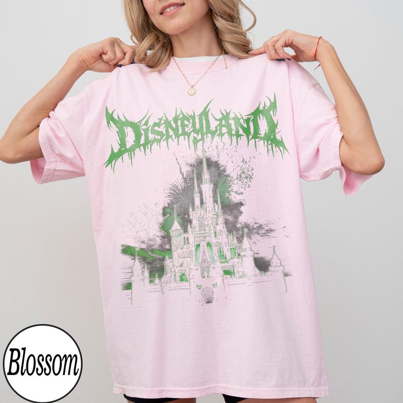 May include: Pink t-shirt with a green and white graphic of a castle and the word "Disneyland" in a spiky font. The shirt is a light pink color and has a round neckline.